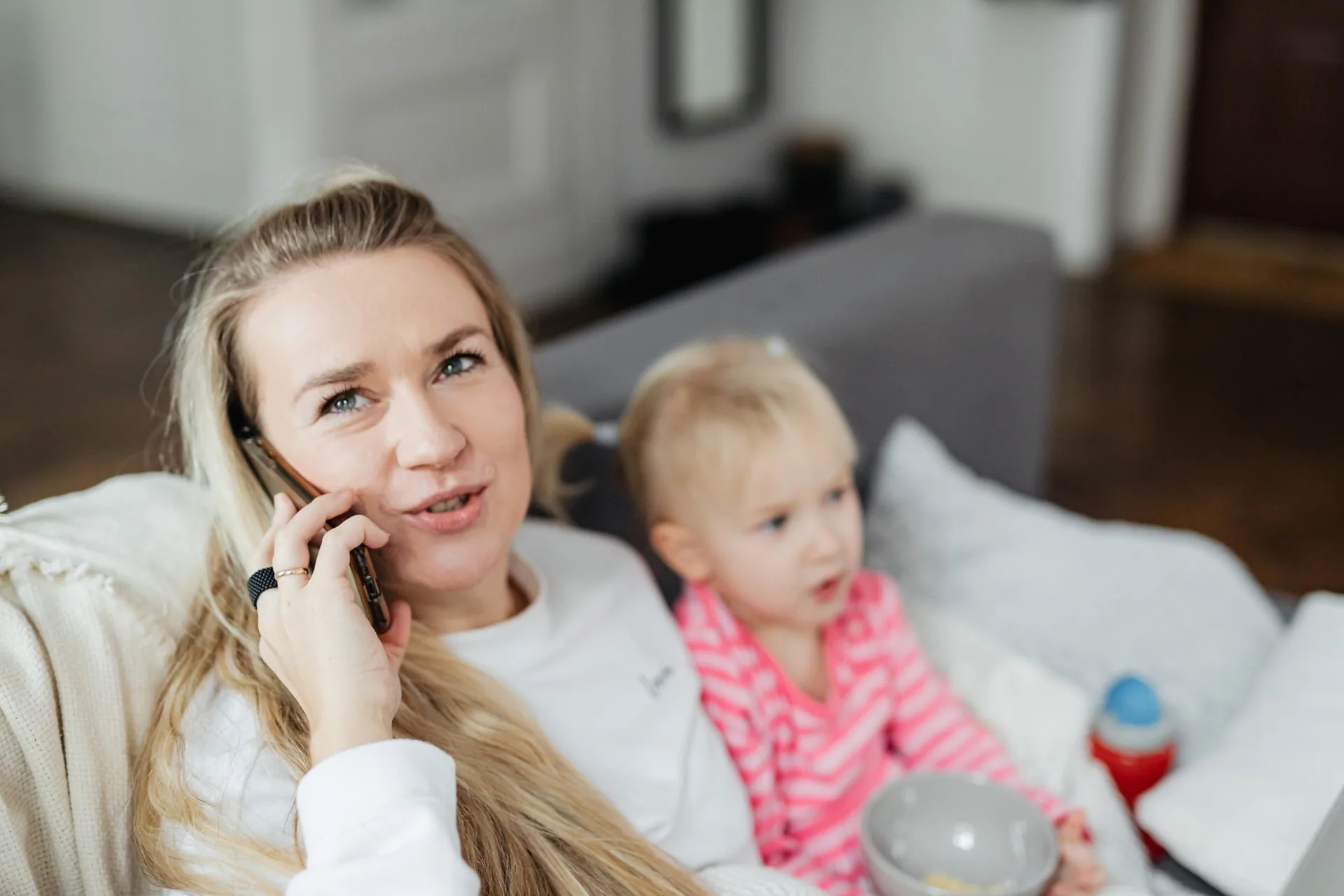 A mom on the phone sitting on a couch with her toddler beside her